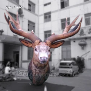 Front view of a giant inflatable reindeer head with realistic brown fur and large antlers, placed on a green mat outside a factory for quality inspection.
